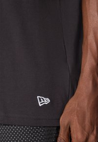 Black t-shirt with short sleeves, cotton material, featuring a small white embroidered logo at the hem. Close-up view of fabric texture.