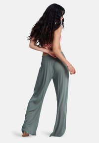 Φαρδιά παντελόνια σε ανοιχτό πράσινο χρώμα, κατασκευασμένα από ελαφρύ, υφή ύφασμα με ελαστική μέση. Το μοντέλο φαίνεται από το πλάι, ξυπόλητο.