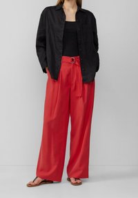 Camisa de lino negra de botones, top negro, pantalones rojos de gran tamaño con cinturón y hebilla circular, y chanclas marrones.