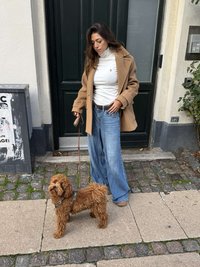 Osoba v béžovém kabátu přes bílý rolák stojí na kamenném chodníku s hnědým kadeřavým psem na vodítku, obklopená budovami.