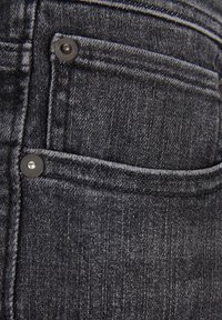 Los pantalones vaqueros de mezclilla oscura cuentan con un bolsillo trasero, botones de metal y una sutileza en la textura del bordado, enfatizando un diseño elegante y minimalista.