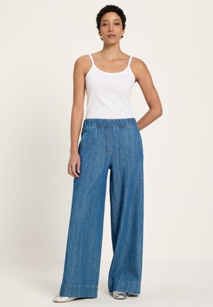 Woman wearing white spaghetti strap top, loose high-waist blue denim pants, and silver shoes, posing against a plain light background.