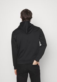 Uomo con capelli castani che indossa una felpa nera e pantaloni neri, in piedi con una mano nella tasca posteriore su uno sfondo bianco semplice.