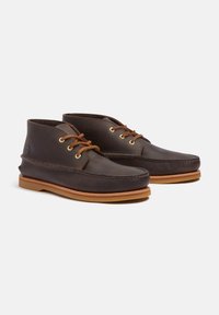 Bocanci chukka din piele maro închis cu ochiuri aurii, detalii cusute și o talpă din cauciuc maro deschis. Au un design cu vârful rotund.