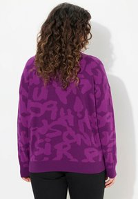 Pull violet avec un motif texturé audacieux en violet plus foncé. Il présente une coupe décontractée et un ourlet côtelé. Porté par-dessus un pantalon noir.
