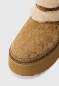 Bota de ante marrón con patrón circular embosado, cuello de borreguito y suela gruesa en contraste. Superficie texturizada con un brillo sutil.