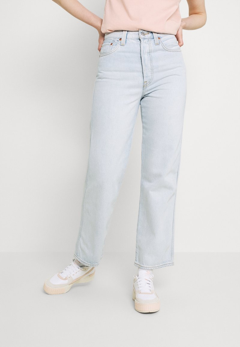 Henkilö, jolla on päällään vaaleansinisiä suoraan leikattuja farkkuja, vaaleanpunainen paita, valkoiset sukat ja valkoiset tennarit, joissa on beige pohjat, seisoo kädet lanteilla.