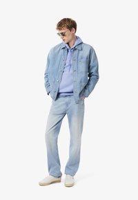 Chaqueta de mezclilla azul claro sobre una sudadera lila, combinada con jeans azul claro y zapatillas blancas. Las gafas de sol añaden un detalle elegante.