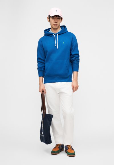 Άνδρας που φορά μπλε φούτερ με κουκούλα, άσπρο παντελόνι, ροζ καπέλο, κρατώντας μια μπλε τσάντα ώμου με καφέ λουριά, στέκεται σε λευκό φόντο.