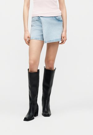 Naine, kes kannab helehalli asümmeetrilist teksamini seelikut, kahvaturoosat varrukateta topi ja musti põlvikuga kõrge kontsaga saapaid.