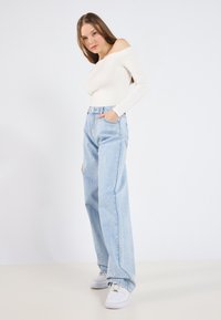 Haut blanc à épaules dénudées avec des manches longues, associé à un jean taille haute large bleu clair, complété par des baskets blanches sur un sol neutre.