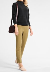 Black textured pullover, olive tapered trousers, and light pink slip-on shoes. A small burgundy shoulder bag hangs from the arm.