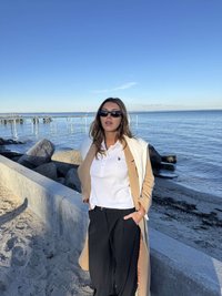 Beżowy długi płaszcz na białej koszuli z kołnierzykiem i czarnych spodniach. Okulary przeciwsłoneczne, falowane włosy, stojąca na piaszczystej plaży przy wodzie.