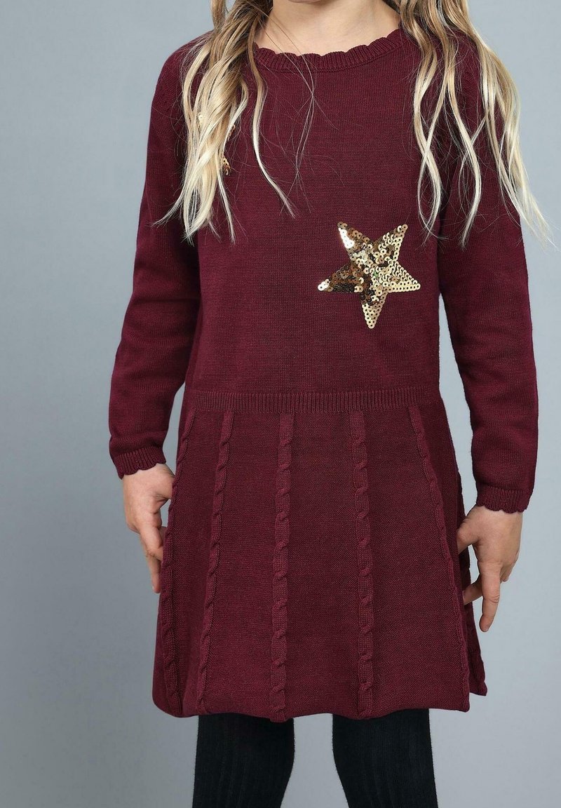 Enfant portant une robe en maille bordeaux à manches longues avec des motifs torsadés et une étoile en sequins dorés sur la poitrine, sur fond gris.