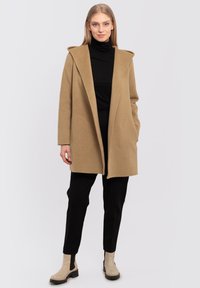 Camel-colored coat with a wide hood, long sleeves, and open front design. Paired with black pants and beige ankle boots. Smooth texture.