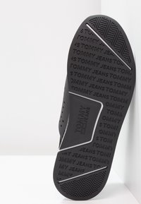 Czarna gumowa podeszwa sandałów z wytłoczonym logo „TOMMY JEANS”, teksturowana dla lepszej przyczepności, z kontrastującym białym akcentem i perforacjami po bokach.