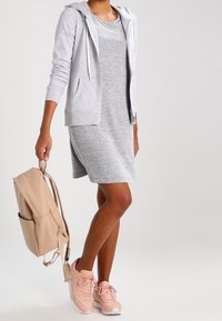 Szara sukienka o swobodnym kroju, zestawiona z jasnoszarą bluzą na zamek, różowymi trampkami i beżowym plecakiem. Gładkie tekstury i luźny design.