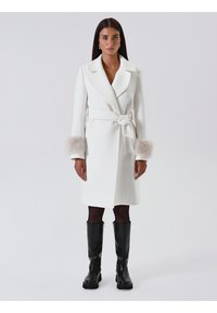 Weiße Wickeljacke mit Bindegürtel, ausgestattet mit leichten Pelz-Akzenten an den Bündchen. Glatte Textur, lange Ärmel und knielanges Design. Schwarze kniehohe Stiefel.