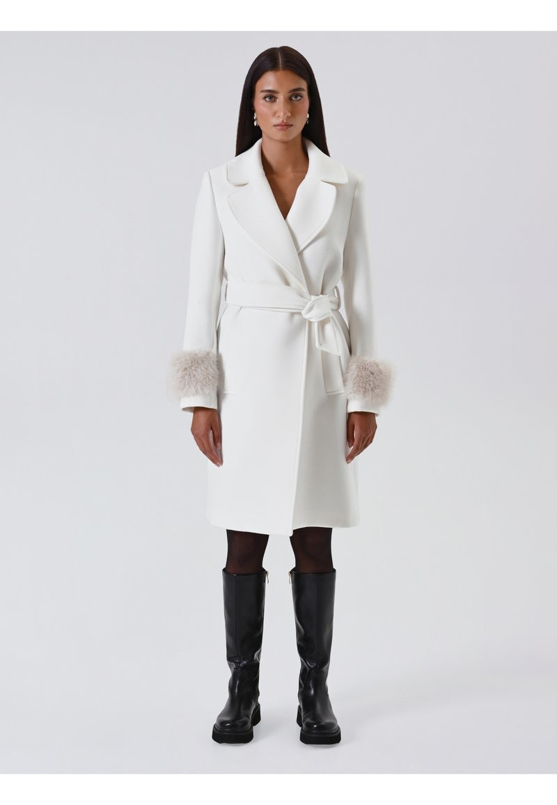 Weiße Wickeljacke mit Bindegürtel, ausgestattet mit leichten Pelz-Akzenten an den Bündchen. Glatte Textur, lange Ärmel und knielanges Design. Schwarze kniehohe Stiefel.