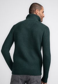Donkergroene gebreide trui met hoge kraag, een textuurpatroon en geribbelde randen. Zichtbare spikkels door de stof heen.