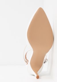 Talons aiguilles blancs avec un bout pointu, un dessus texturé et une semelle beige comportant un petit accent orange à la pointe. Sangle de cheville ajustable incluse.