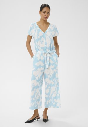 Žena nosi svetlomodrý a biely kvetinový krátky overal s širokými nohavicami, prednými gombíkmi, opaskom na zaväzovanie, rukami v vreckách a čiernymi sandálmi s otvorenou pätou.