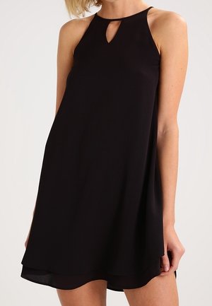 Ärmelloses schwarzes A-Linien-Kleid mit Neckholder und kleinem Schlüssellochausschnitt an der Brust, getragen von einer stehenden Person.