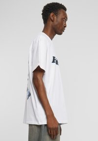 Hvid bomulds T-shirt med korte ærmer, rund halsudskæring og et blåt grafisk tryk på ryggen. Løs pasform med lige kant.