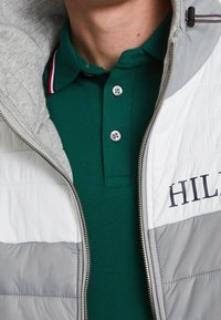 Grijze en witte gewatteerde jas met een rits, gelaagd over een groene poloshirt. De poloshirt heeft een textuurstof en een knoopsluiting met drie knopen.