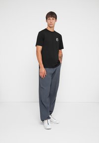 Sort bomulds T-shirt med korte ærmer og rund halsudskæring, parret med løse, grå bukser og hvide sneakers. Diskret grafisk detalje på trøjen.