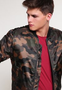 Maskēšanas bumbierim līdzīgs žakets zaļā, brūnā un melnā krāsā. Tam ir ribota apkakle, rāvējslēdzējs un sānu kabatas. Gluda, viegla auduma.