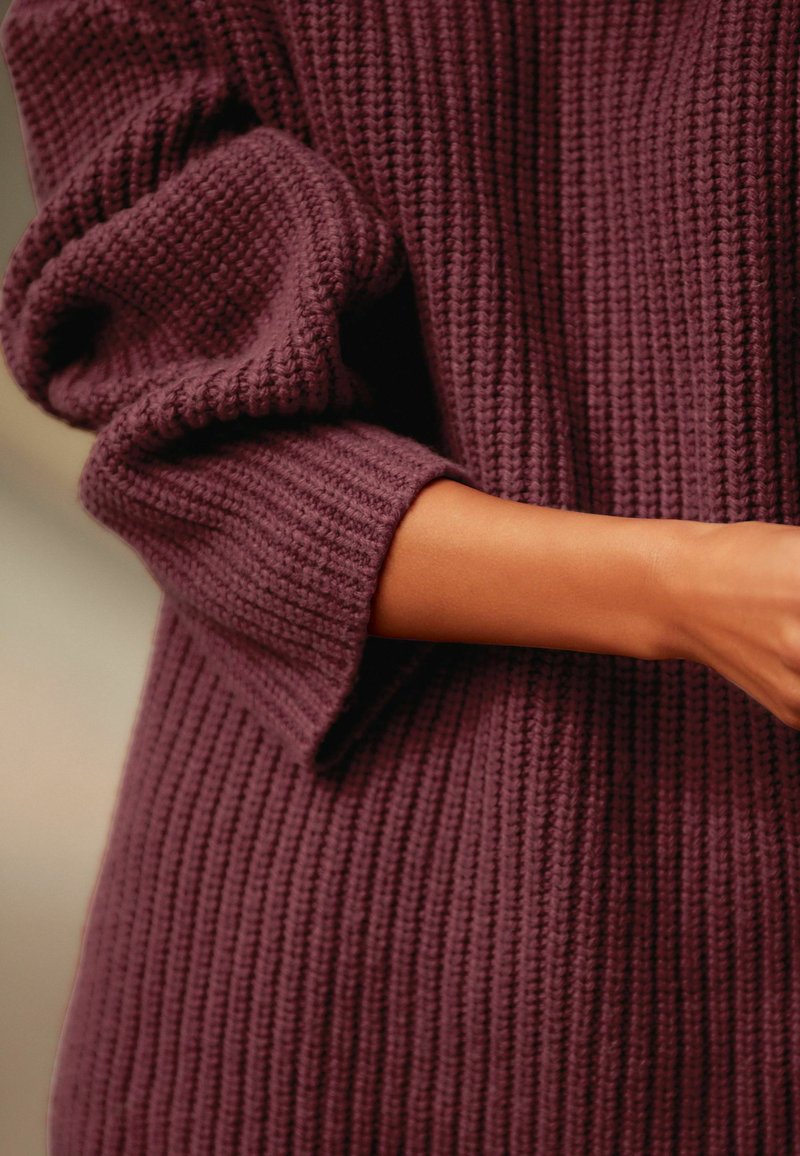 Burgunderfarbener Strickpullover mit geripptem Textur, oversized Ärmeln und lockerer Passform. Sichtbarer Arm zeigt glatte Haut gegen den Stoff.