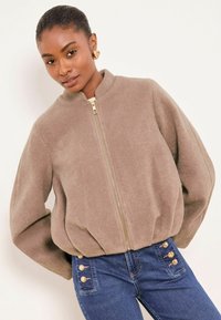 Chaqueta bomber marrón claro con cremallera y cuello alto; tejido suave y texturizado con mangas voluminosas. Los vaqueros presentan detalles con botones dorados.