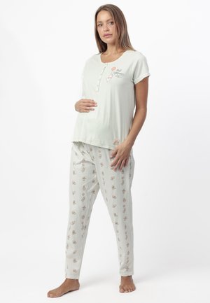 Mujer embarazada con camiseta blanca de maternidad abotonada con el texto "Mejor Mamá" y pantalones de pijama estampados, de pie descalza sobre fondo blanco.