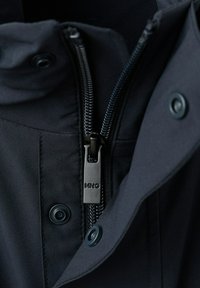 Gros plan sur une veste noire montrant une fermeture éclair centrale avec un tirette gravé "MNG" et des boutons-pression sur la patte en tissu.