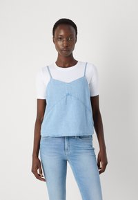 Denim camisole in light blue, featuring thin straps and a triangular bib design. Paired with a fitted white long-sleeve shirt and blue jeans.