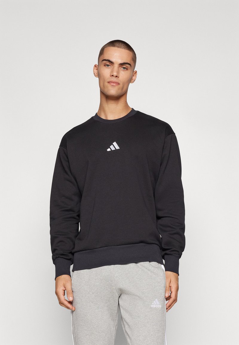 Sort sweatshirt med ribbet rund hals, lange ærmer og lille hvid Adidas-logo på brystet, parret med lysegrå sweatpants.