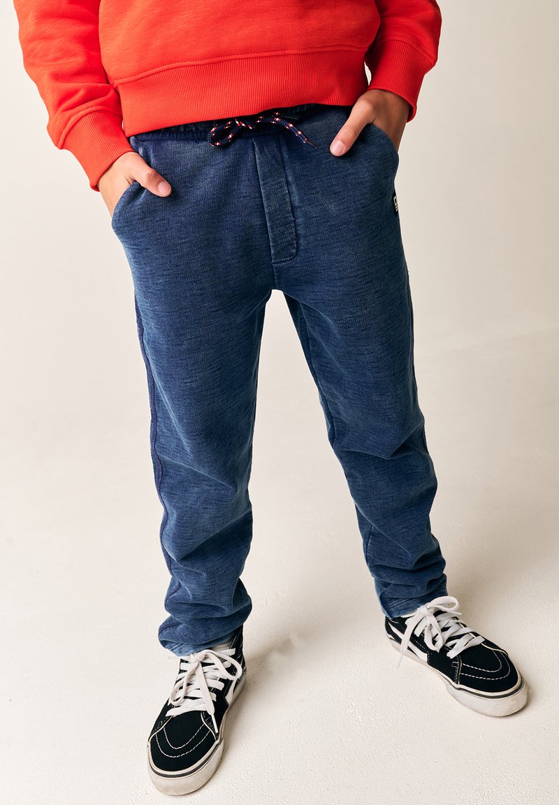 Mørkeblå joggerbukser med en struktureret overflade, elastisk talje og snor. Parret med sorte sneakers og hvide snørebånd.