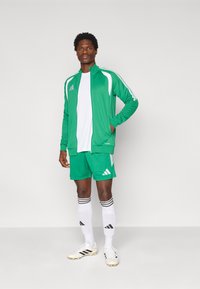 Πράσινο αθλητικό μπουφάν με λευκές λεπτομέρειες, συνδυασμένο με αντίστοιχες πράσινες βερμούδες, λευκές κάλτσες και ανοιχτόχρωμα ποδοσφαιρικά παπούτσια με μαύρες ρίγες.