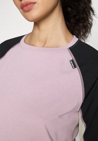 Lys rosa langermet skjorte med svarte raglanermer, preget av kontrasterende sømmer og en liten logoetikett på øvre høyre skulder.