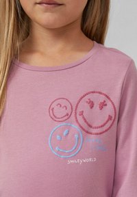 Ein Kind trägt ein pinkes Shirt mit drei Pailletten-Smiley-Gesichtern in Pink und Blau sowie dem Text "Teile ein Lächeln" und "Smileyworld."