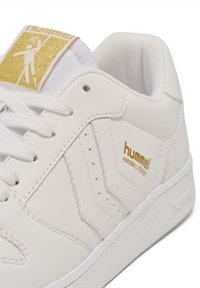White leather sneaker with gold accents, featuring a textured toe cap, perforated side design, and white rubber sole. Logo on tongue.