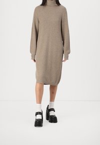 Robe en maille beige à col roulé avec manches longues, arrivant mi-mollet. Associée à des chaussures à plateforme noires et des chaussettes blanches remontant à la cheville pour créer un contraste.