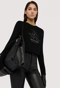 Svart tröja med logotydelse, kombinerad med svarta läderbyxor. En stor svart läderväska vilar i modellens hand, med stilren hårdvara.