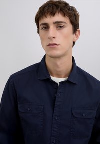 Marineblaue, knöpfbare Baumwollbluse mit zwei Vorderentaschen und Kragen. Glatte Textur ohne sichtbare Muster oder Akzente.