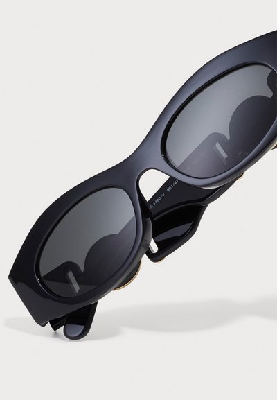 Lunettes de soleil noires surdimensionnées avec une finition brillante, lentilles ovales et montures angulaires distinctes. Des accents dorés rehaussent le design.