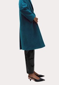 Abrigo color verde azulado de tejido texturizado, con silueta larga y frente abierto, combinado con pantalones negros brillantes y tacones slingback negros puntiagudos.