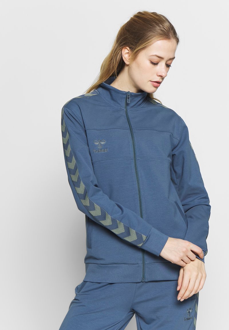 Niebieska kurtka zapinana na zamek z wysokim kołnierzem, z kontrastowymi wzorami w kształcie zygzaka na rękawach oraz logo na piersi. Miękka faktura materiału.