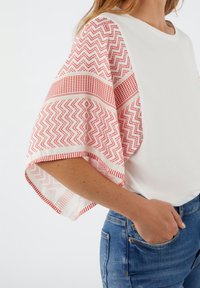 Hvid top med brede røde mønstrede ærmer med zigzag- og gitterdesign. Parret med blå denimjeans. Blød tekstur, afslappet pasform.