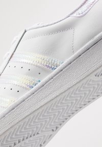 White leather sneaker with holographic stripes, textured sole, and perforations. Embossed "SUPERSTAR" logo on side. Overall sleek design.